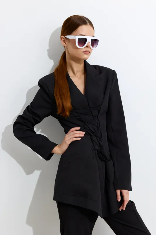Fashion woman wearing oversized white sunglasses and a black tailored jacket, posing confidently against a bright white minimalist background with sleek hairstyle.