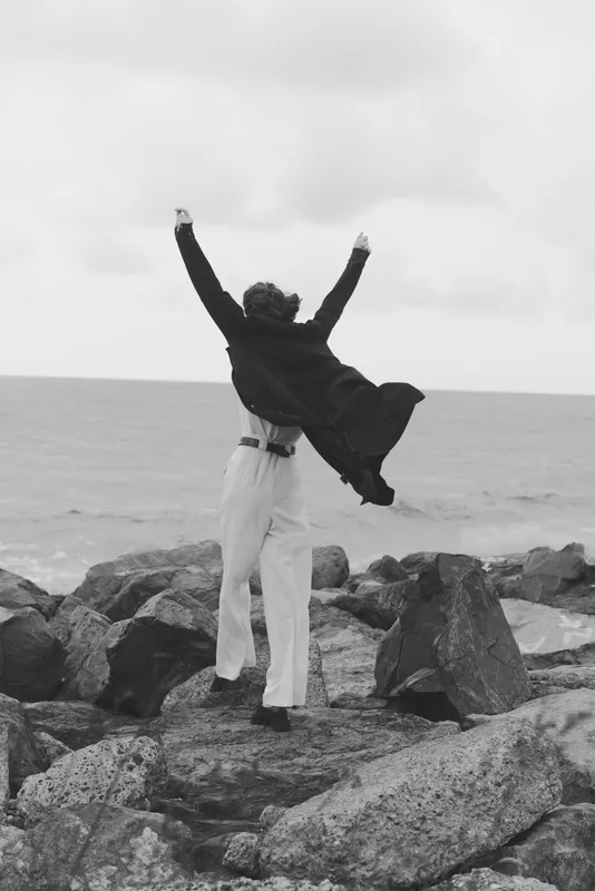 Individual stands on rugged rocks beside the sea, arms raised in triumph, black and white scene capturing freedom, celebration, and outdoor energy amid wind and coast, conveying motivation,