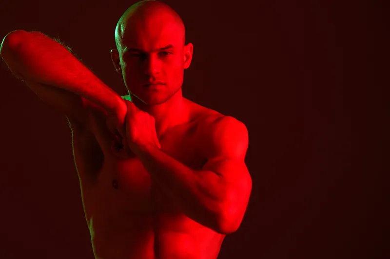 Sculpted man posing dramatically in a low light setup, red and dark color palette emphasizing muscle tone and defined features, perfect for fitness themes.