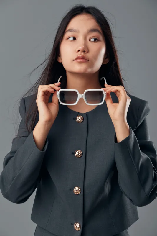 Young woman holding white sunglasses dressed in professional dark blazer with buttons posing on gray background. Elegant girl with long hair looking aside in studio portrait photography.
