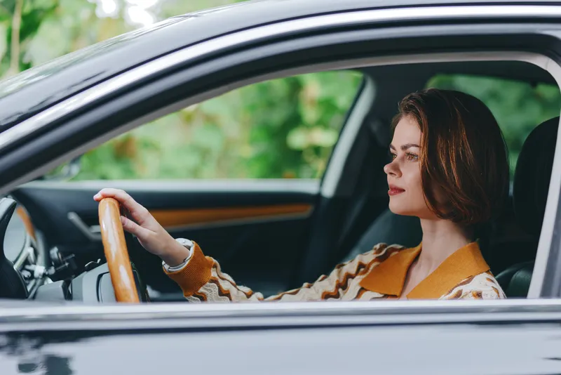 A focused woman drives a car, hands on the wheel, eyes ahead, wearing a warm sweater, with a calm interior and soft natural light highlighting concentration and independence.