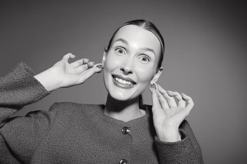 Happy Caucasian woman with fair skin smiles while adjusting earrings in studio with plain background. Concept of beauty, fashion, and happiness.