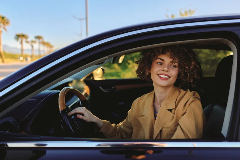 A woman driving a car with a bright smile, casual jacket, retro film color tone. A lifestyle portrait captured in a candid moment, conveying optimism, independence, and everyday joy.