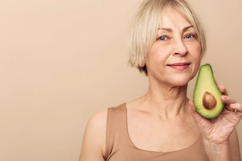 Smiling mature woman with short blonde hair holds fresh avocado half near face on beige background. Concept of healthy lifestyle, nutrition, natural beauty, and wellness for older adults.