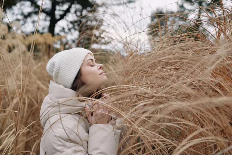 Woman wearing a white winter coat and knit hat stands among tall grass, eyes closed, savoring serene nature and a gentle cold breeze in tranquil outdoor light.