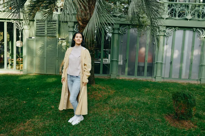 Woman in a long beige coat standing on a grassy lawn outdoors, casual fashion, daytime light, lush garden backdrop and modern glass facade, relaxed pose in natural setting, serene atmosphere