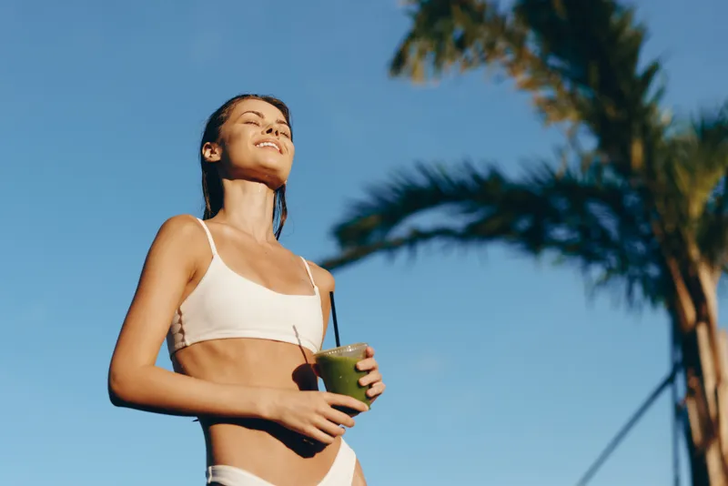 Cheerful woman outdoors, sunlit moment, white bikini, sipping a green drink, warm breeze, palm trees in background, relaxed posture, natural colors, vibrant atmosphere, healthy lifestyle vibe