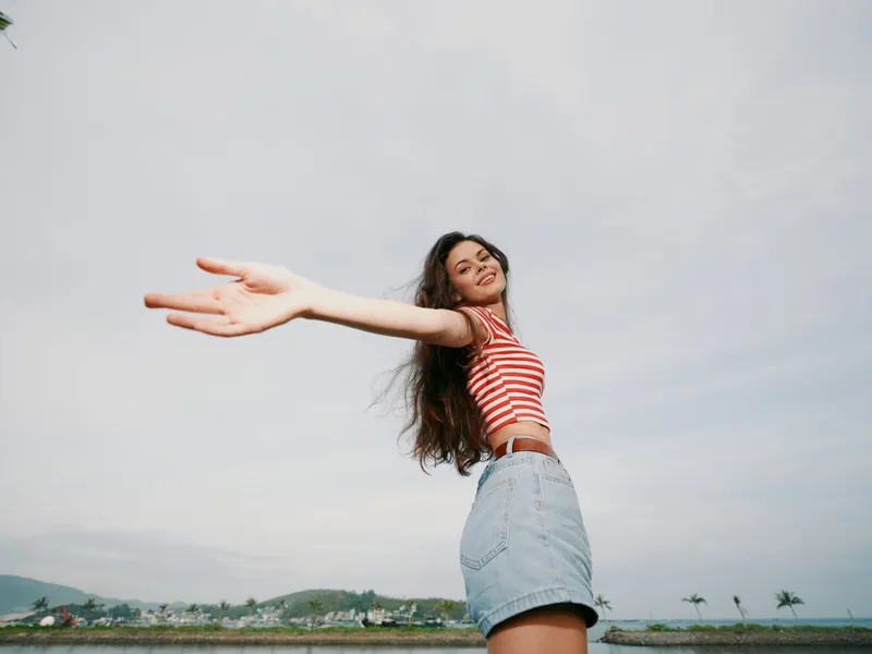 Young woman with outstretched arms stands outdoors, smiling toward the camera, wearing a red striped crop top and denim skirt, wind in her hair under a bright sky.