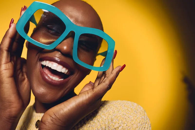 Happy black woman with dark skin wearing large blue glasses smiles joyfully on yellow background. Close-up portrait shows positive mood, fashion, and beauty concept with vibrant colors.