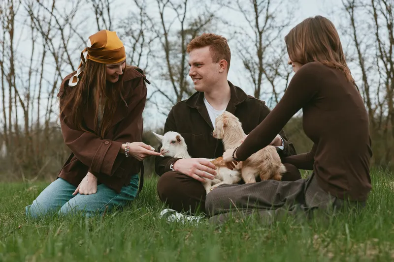 Three friends outdoors on a meadow share a moment with a goat kid, cuddling and smiling while the scene conveys a warm lifestyle, natural light, candid connection, gentle play, and a carefree mood.