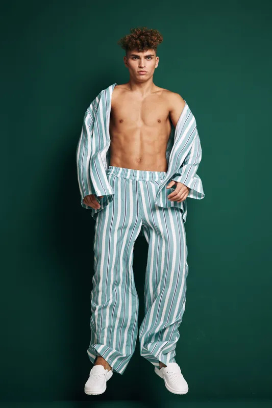 Young man with curly hair wearing striped pajamas posing on green background. Shirt open, bare chest and athletic body, comfortable cotton clothing and stylish sleepwear model.
