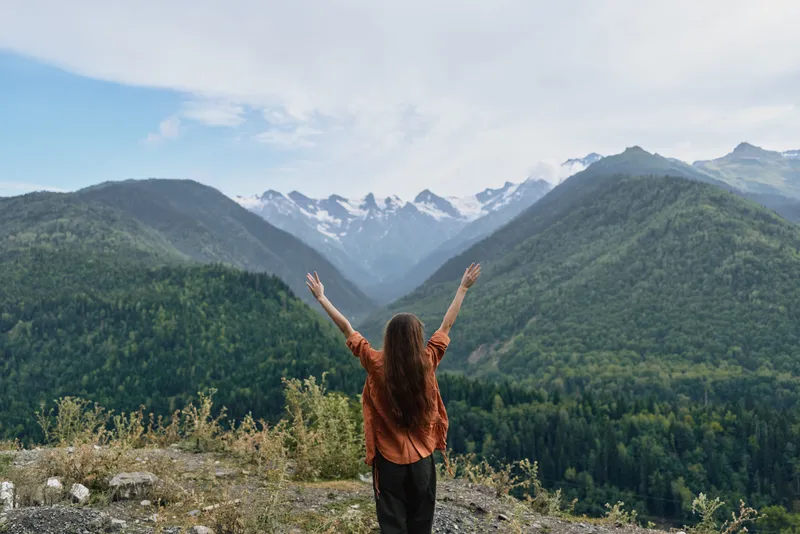 Woman stands with arms raised at a mountain overlook, savoring a vast landscape of forests and distant peaks under a clear sky, conveying freedom and adventure