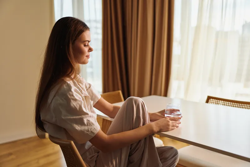 woman holding glass of water at table in home morning light, sitting on chair and relaxed, thoughtful portrait in cozy loungewear by window with soft curtains and sunlight