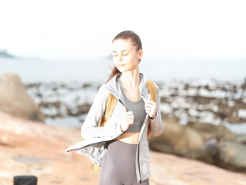 Young fitness woman wearing a gray workout jacket and yoga pants, enjoying the sun by the beach, promoting healthy lifestyle and outdoor activities