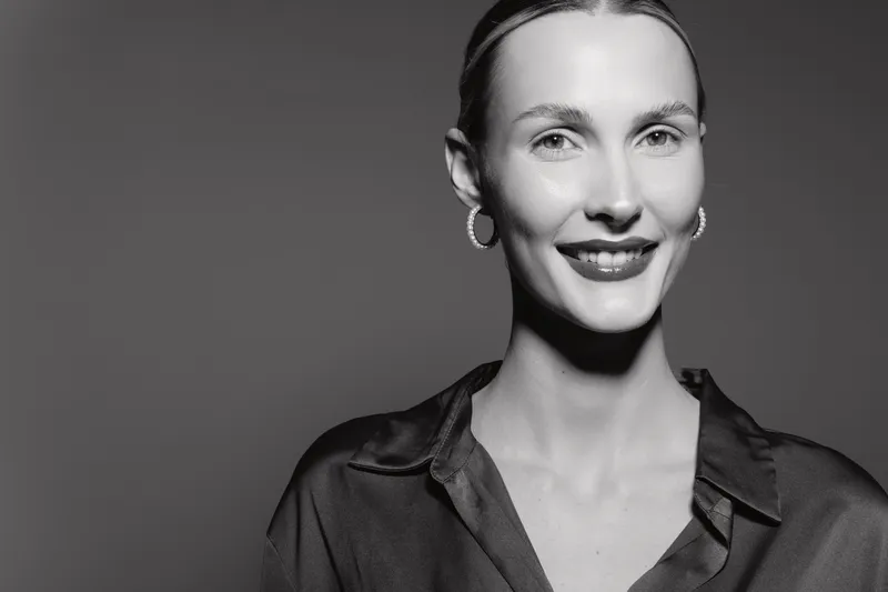 Smiling Caucasian woman with fair skin and slicked back hair wears dark blouse and hoop earrings, posing against plain background. Studio portrait.