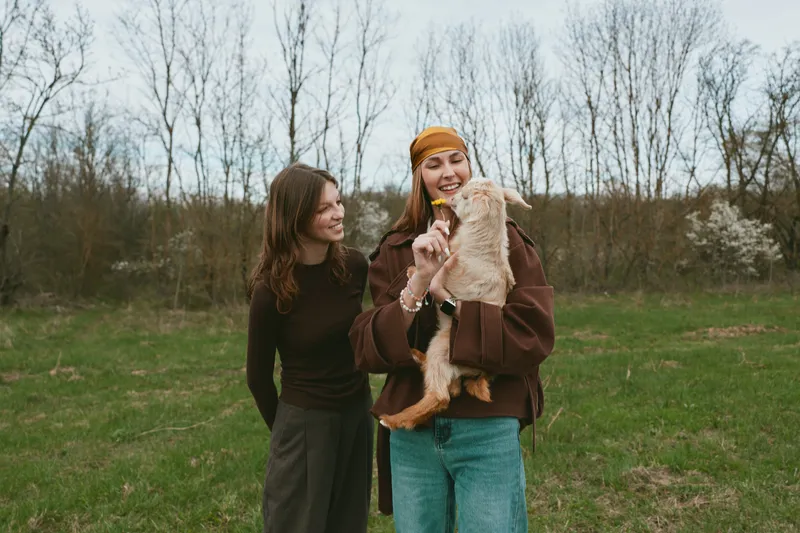Two women share a sunny outdoor moment with a joyful goat kid in a rural field, capturing candid friendship, nature, and simple countryside happiness in daily life.