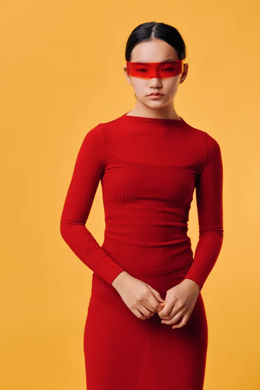 Woman in red dress and red transparent glasses posing with hands clasped against yellow background. Modern fashion style with minimal expression and clean studio lighting for portrait.