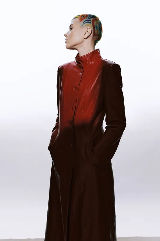 woman with colorful short hair wearing a long brown leather coat standing with hands in pockets, stylish outfit, clean white background, bold fashion statement