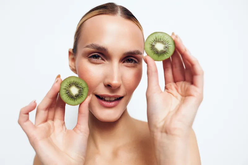 Healthy woman holding kiwi slice near face, smiling in closeup on clean white background, beauty skincare and freshness concept, green fruit in hands, copy space