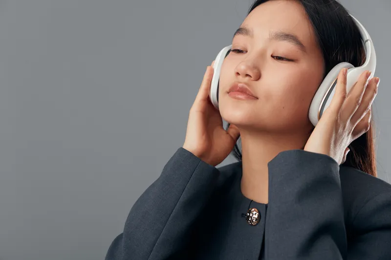 Young woman with closed eyes wearing white headphones and dark jacket listening to music or audio content in calm, relaxed pose on neutral gray background in studio.