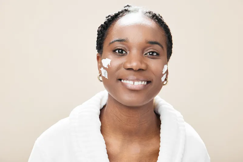 Smiling Black woman applying face cream, promoting skincare and self care with a neutral background, ideal for wellness and beauty concepts