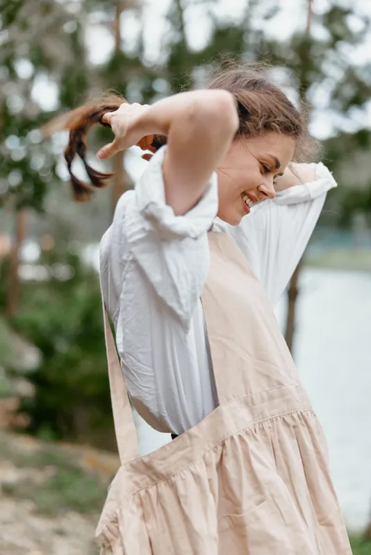 Joyful young woman in a light summer dress enjoying nature by the river, exhibiting happiness and carefree emotions with greenery in the background