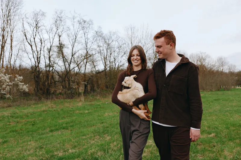 A smiling couple walks in a grassy field while holding a friendly goat kid, enjoying a relaxed outdoor moment. The scene captures lifestyle, family, and countryside joy, with natural light and a