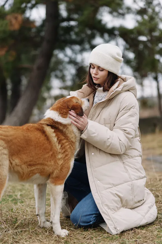 Dog and woman kneeling in a park, outdoor moment with a warm coat and beanie, gentle interaction as she pets the dog.