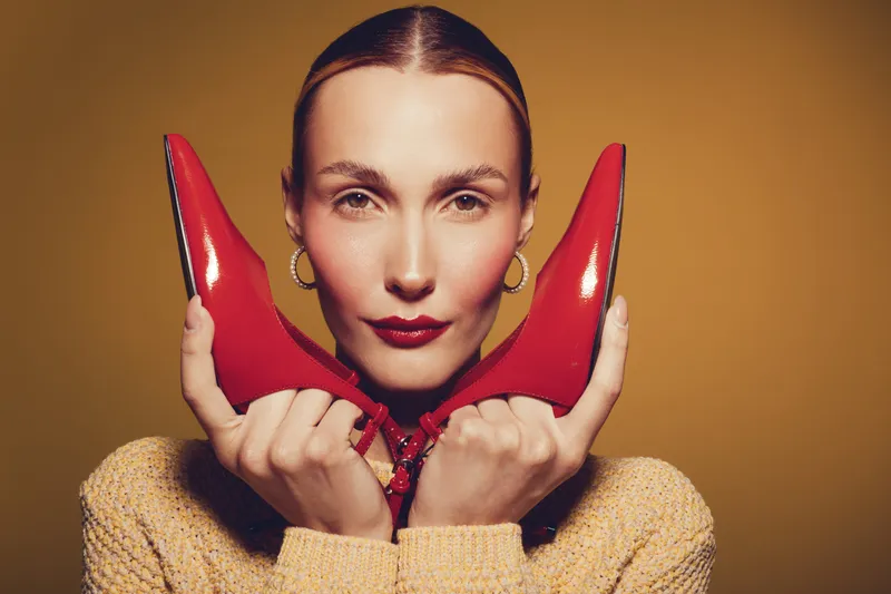 Young woman with slicked hair holds red high heels near face, wearing beige sweater and gold hoop earrings. Studio portrait on warm brown background. Concept of fashion, style, and elegance.