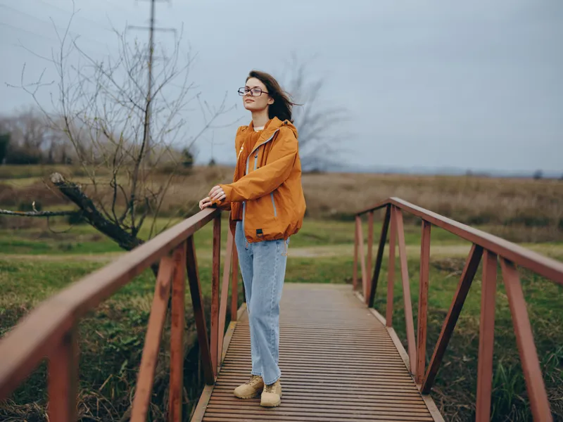 Woman lifestyle on a wooden bridge outdoors, autumn field and bare trees, orange jacket and blue jeans, walking calmly in cloudy daylight, natural portrait, soft wind with gentle expression