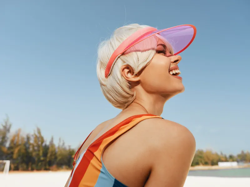 Young woman wearing a bright pink visor and colorful swimsuit smiles against a clear blue sky, embodying carefree summer energy and vibrant outdoor lifestyle.