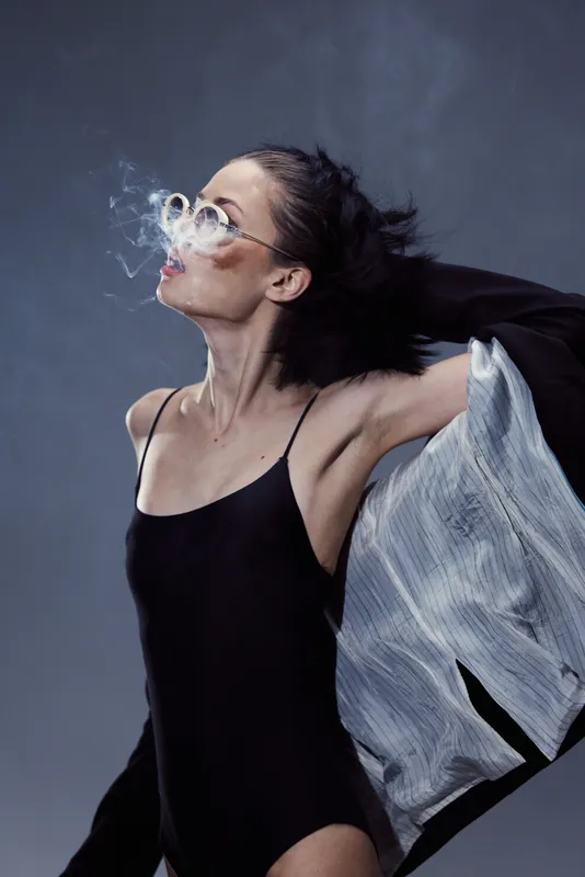 Woman blowing smoke rings wearing black bodysuit and sunglasses with flowing jacket on gray background