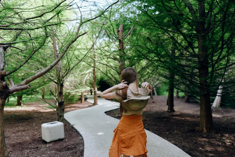Woman walking on winding path through forest trees, back view with hat and orange skirt, outdoors scene of summer park trail, scenic nature stroll and relaxed leisure.