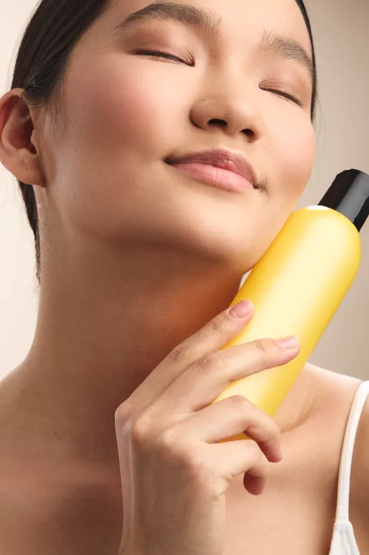Asian woman enjoys a refreshing moment with skincare spray, basking in soft light and a calm smile, conveying wellness, confidence, and modern self care in a serene environment.