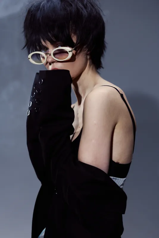 Stylish woman with short dark hair wearing oversized glasses and a black jacket with exposed shoulders in a moody indoor setting