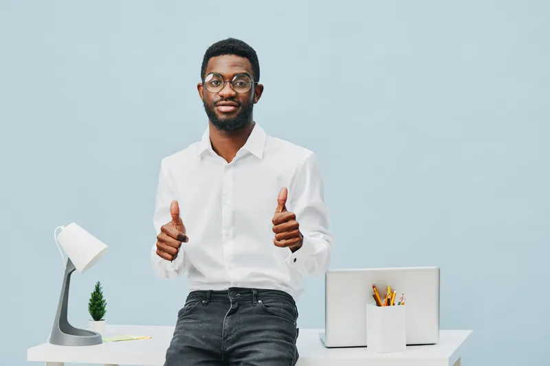 Smiling professional young man of African descent in white shirt standing at modern office desk, giving thumbs up gesture, confident expression, minimalistic workspace with desk lamp and office suppli