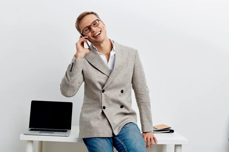 Smiling woman in business casual attire talking on mobile phone in office setting with laptop and folders, looking confident and cheerful, isolated on plain white background. People lifestyle concept
