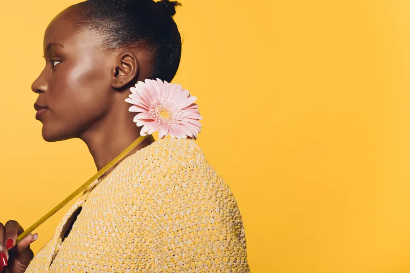 Profile of a young black woman with dark skin holding a pink flower on her shoulder against a yellow background. Concept of beauty, nature, and elegance in studio portrait.