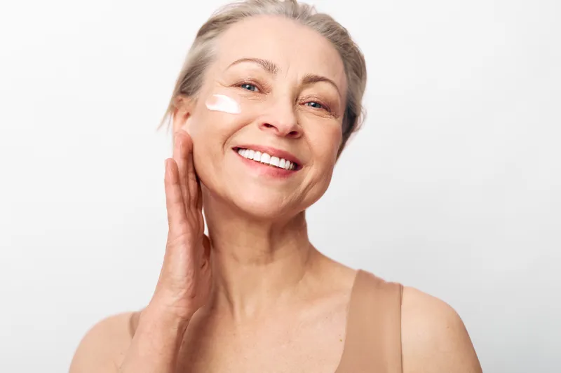 Smiling mature woman applies skincare cream on face with hand touching cheek. Bright white background highlights healthy skin and self care concept for beauty and wellness routine.