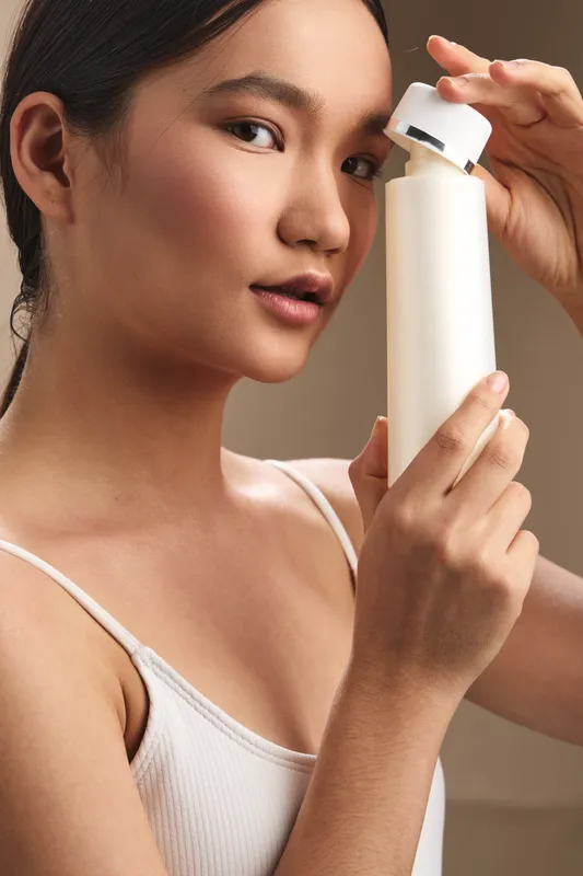 Asian woman holding a sleek bottle in a soft studio setting, preparing to apply product. She gazes gently at the camera with calm confidence, conveying beauty care and personal wellness.
