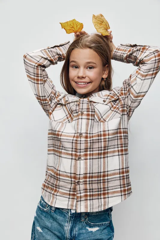 Girl smiling and holding autumn leaves on her head wearing a plaid shirt and denim jeans against white background. Child with casual clothes and fall nature theme.