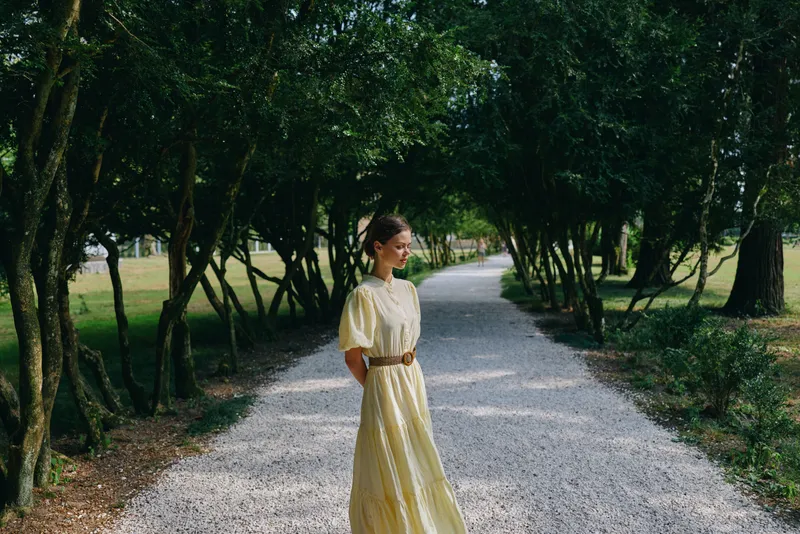 Woman in a yellow dress stands on a gravel path in a lush park, bathed in natural light, creating a serene outdoor portrait with soft shadows and a peaceful mood for photography and lifestyle usage