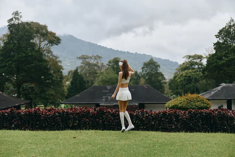 Young woman in a playful white outfit, enjoying nature in a lush green garden, expressing joy, with mountains and a cloudy sky in the background