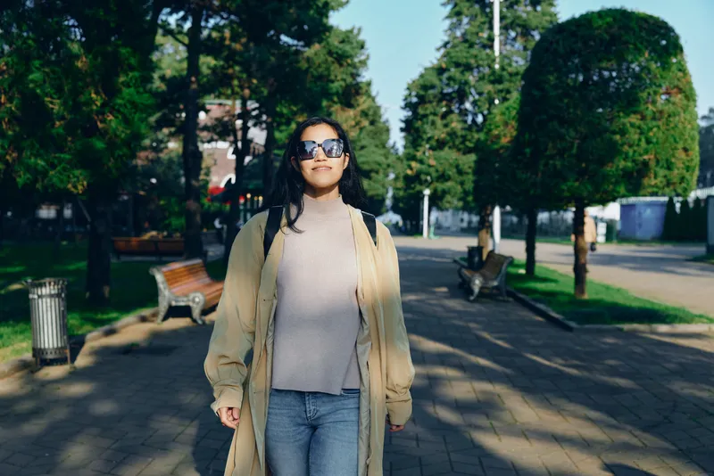 Woman walks along a sunny park street wearing a beige coat and sunglasses, showcasing casual street fashion in an outdoor urban setting with trees, plaza and walkway, conveying relaxed confidence and