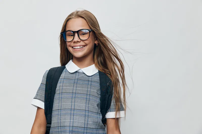 Girl with glasses and long hair smiling wearing school uniform and backpack standing on gray background. Child portrait, education, student with happy expression and casual style.