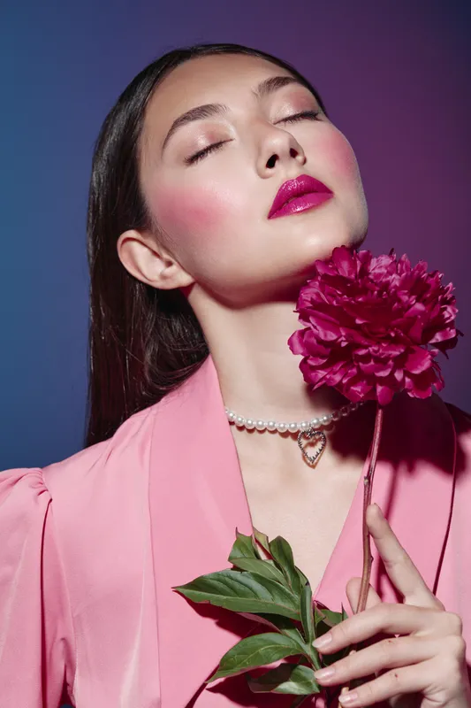 Young woman with closed eyes wears pink blazer and bright pink lipstick while holding vibrant flower near chin. Fashion beauty portrait with creative color styling for cosmetic concept.