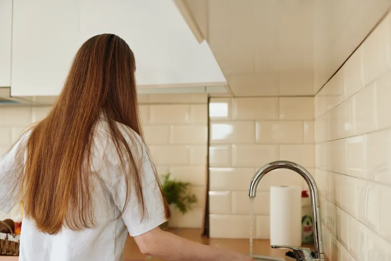 Woman in kitchen at sink in home with long hair cleaning dishes at countertop, casual domestic scene with faucet, tiles and natural lighting showing everyday household routine