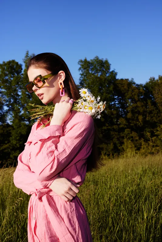 Young woman in a pink dress holding daisies, showcasing a joyful and vibrant summer vibe, set against a lush green field background