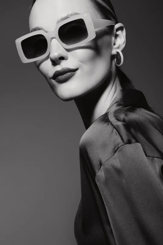 Stylish Caucasian woman with pale skin wearing large sunglasses and hoop earrings poses confidently in a satin blouse. Black and white portrait with dramatic lighting.