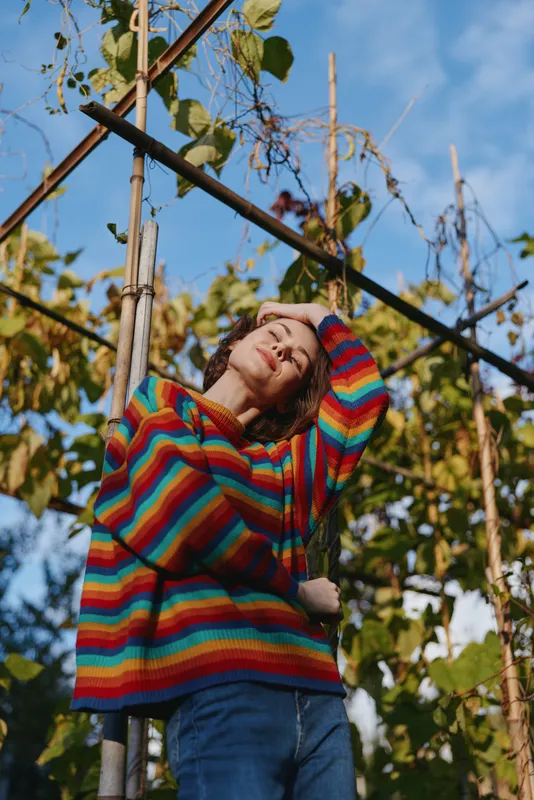 Woman in a colorful striped sweater and jeans stands in a garden by a vine trellis, curly hair and smiling with eyes closed, relaxed casual outfit and warm sunny outdoor lifestyle.